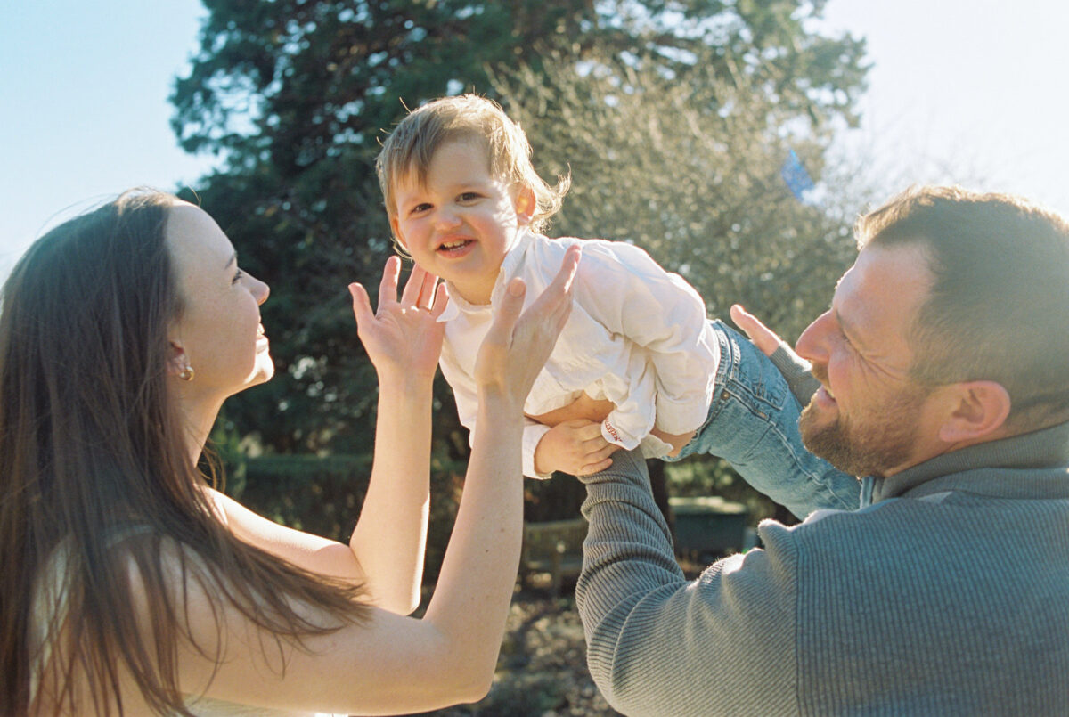 Family photography at Government House in Victoria BC taken on 35mm film. The toddler is being raised in the air like he is flying and his mom is reaching out to touch his face.