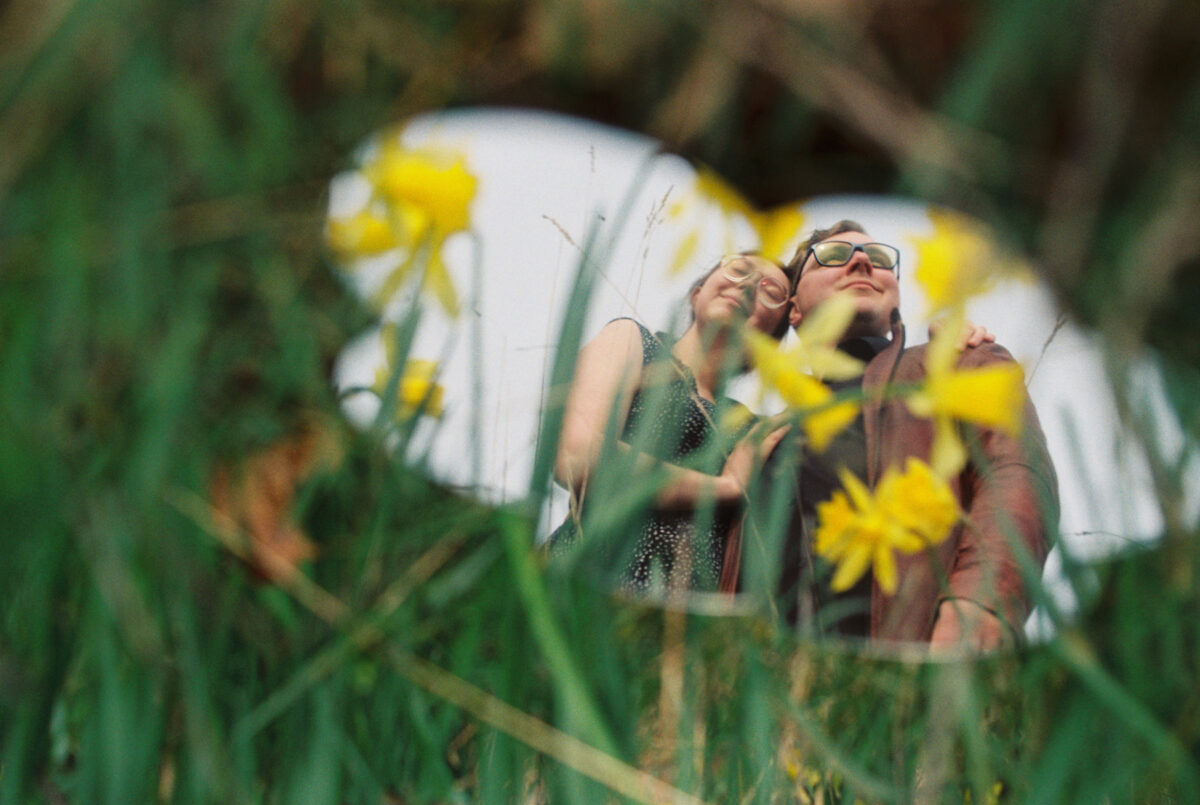 An engagement portrait on Kodak Gold 35mm film in Victoria BC. They are reflected in acrylic glass with grass and daffodils in the foreground. 
