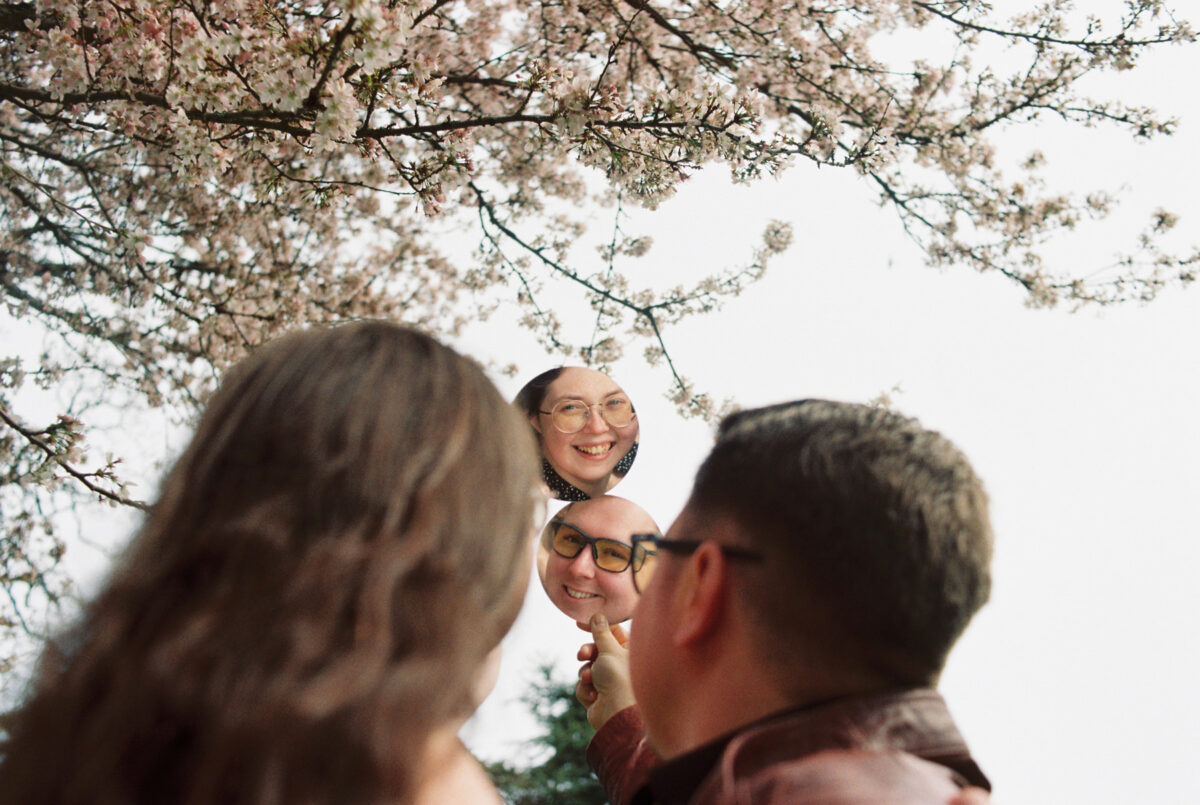 A couple laughs at their reflection in round mirrors held up to cherry blossoms during spring at Beacon Hill Park in Victoria BC. this photo was taken with Kodak Gold 35mm film. 