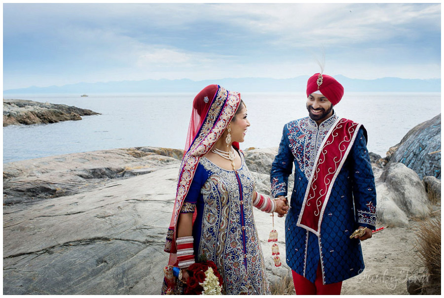 A sikh wedding photography portrait at the mile zero area of Dallas Road in Victoria BC. The sky is blue and the couple is wearing blue and red attire. they are looking at each other and holding hands.