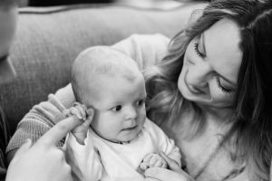 A mother holds her baby during a documentary family photo session at home in Colwood British Columbia near Victoria. The photo is black and white and the baby is poking a finger at her ear.