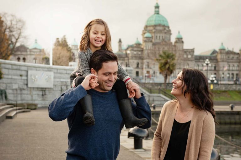 A family of three with a toddler having a fun time with a child on dad's shoulders in front of the BC Legislature buildings in Victoria BC. The photo is in colour and this is a candid family portrait.