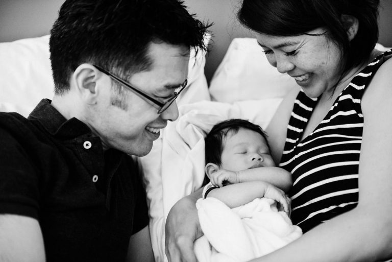 Two parents holding their baby boy while laying in bed in a candid, natural moment during a newborn photography session in Victoria BC