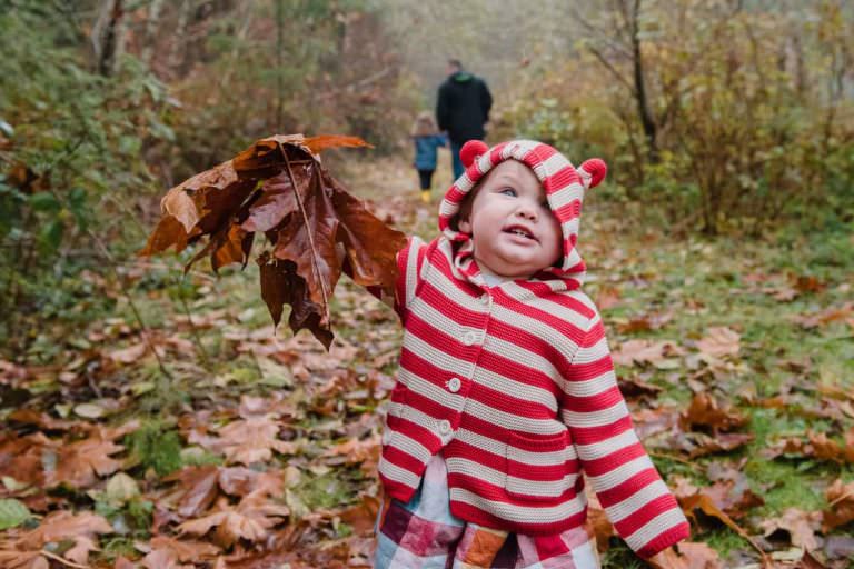 A cute picture of a toddler holding a leaf and wearing a striped sweater hoodie with ears on a fall day in Victoria BC. A colour photograph taken by family photographer Christina Craft
