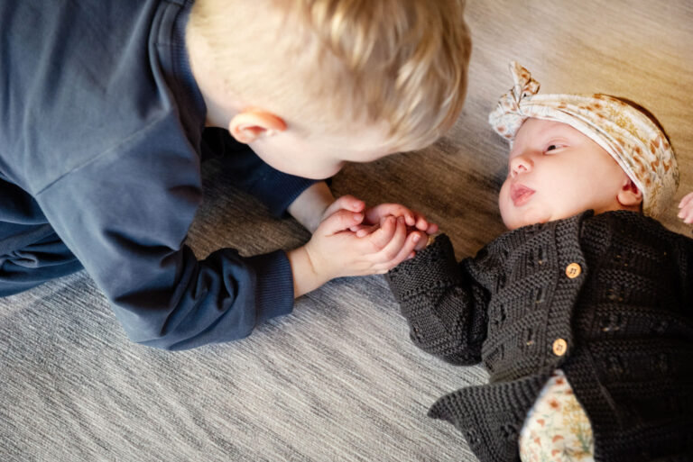 A toddler holds the hand of his newborn baby sister who is laying on the floor at their home during a documentary newborn photo shoot in Victoria