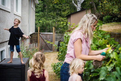 A documentary family photo take in a family's backyard in Victoria BC. The mother and two daughers are picking raspberries while their brother is standing on a container singing.