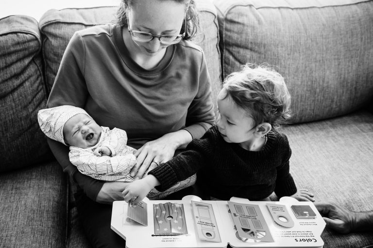 A toddler reads to his newborn baby sister as his mother holds her. He's pointing to a picture book. The photo is in black and white and it's taken in the client's living room in victoria bc during a lifestyle photo session.