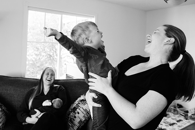 Two moms and their son are having fun in their living room while participating in a documentary family portrait session by FunkyTown Photography.