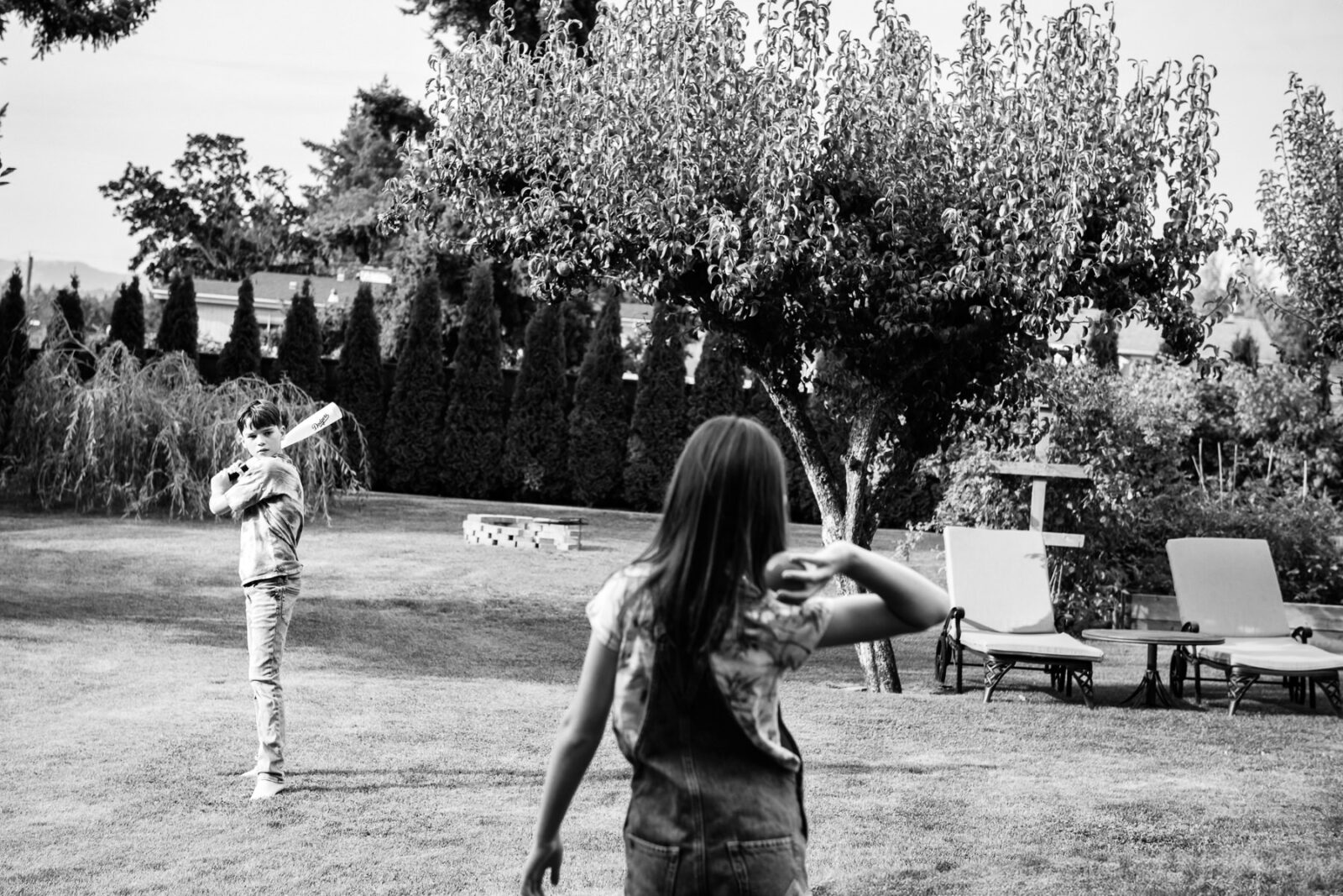 A girl throws a ball towards her brother's bat in their backyard during a candid family photo session in Victoria BC