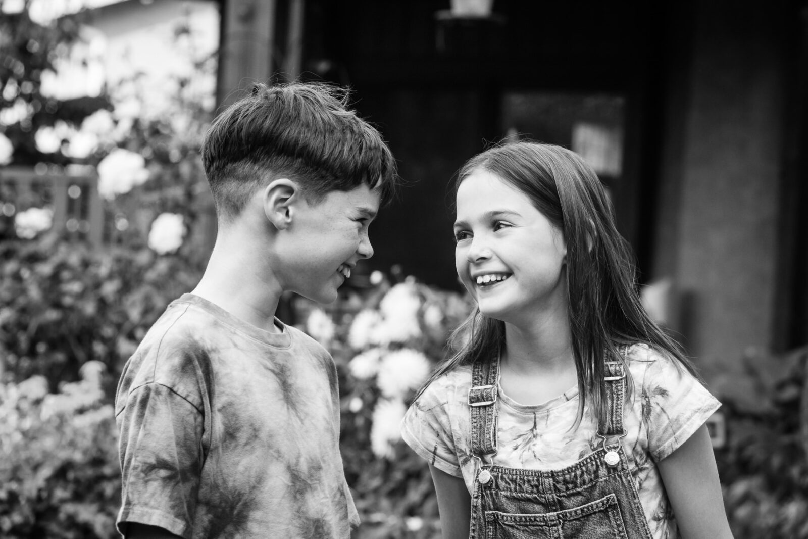 A picture of a brother and sister looking at each other and about to laugh. The photo is a portrait in black and white taken in Victoria BC