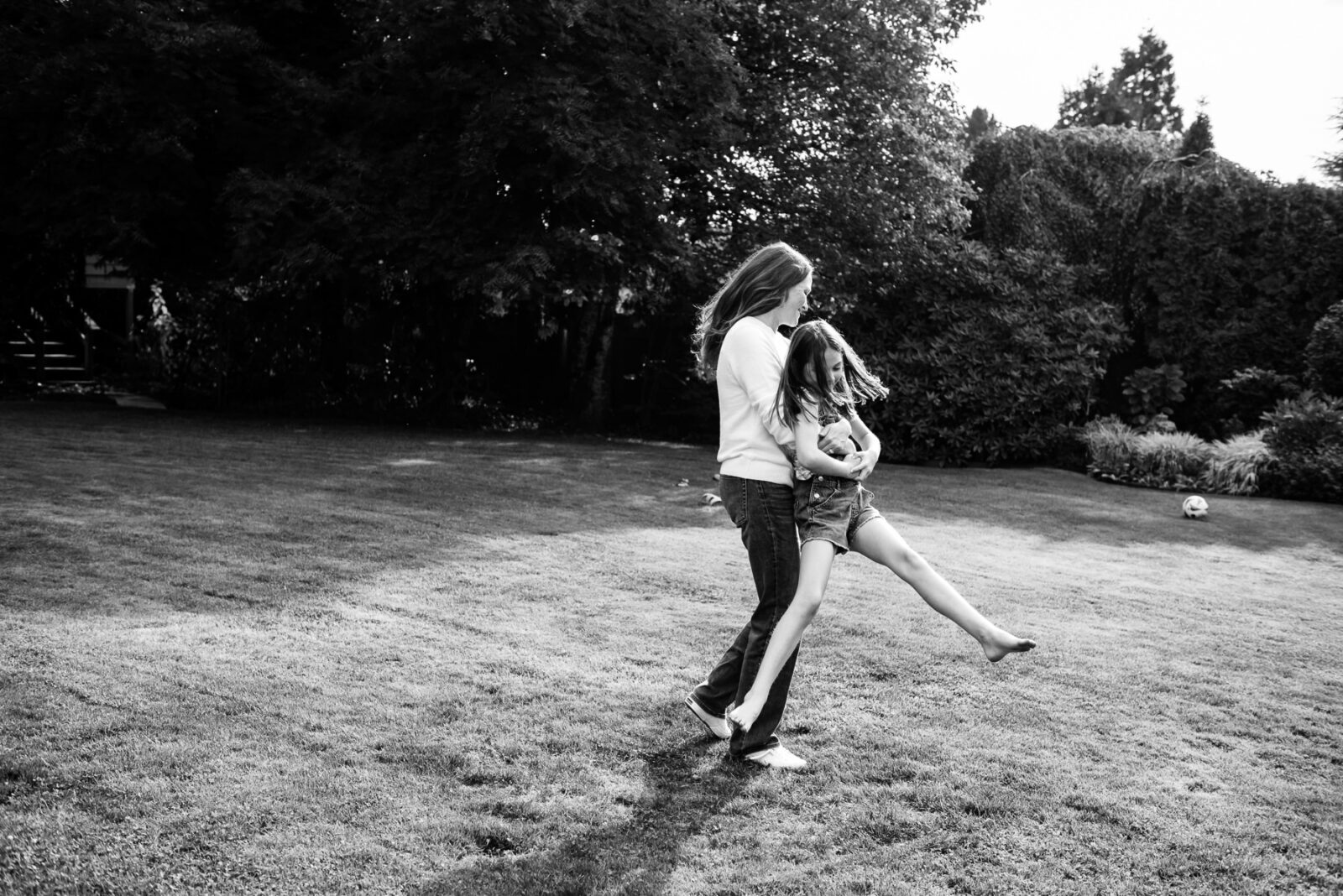 A black and white photo of a mom twirling her daughter around on their lawn in the backyard of their home in Victoria BC