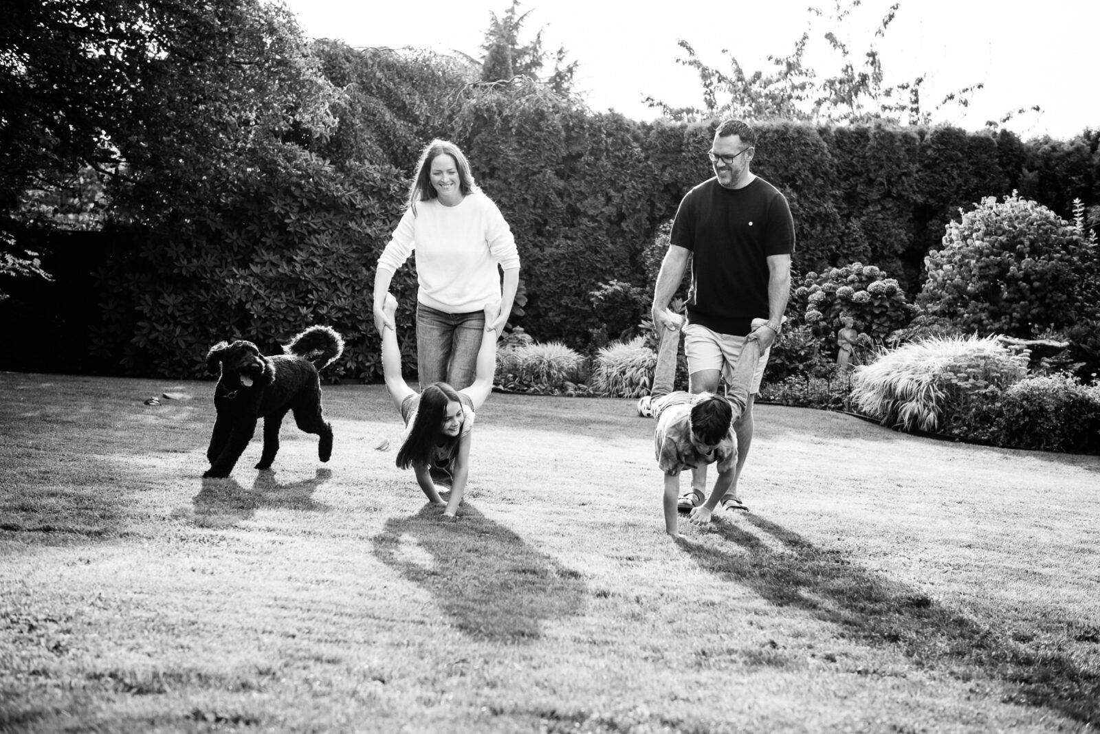 Two parents and two kids play a wheelbarrow game in their backyard as their dog runs around.
