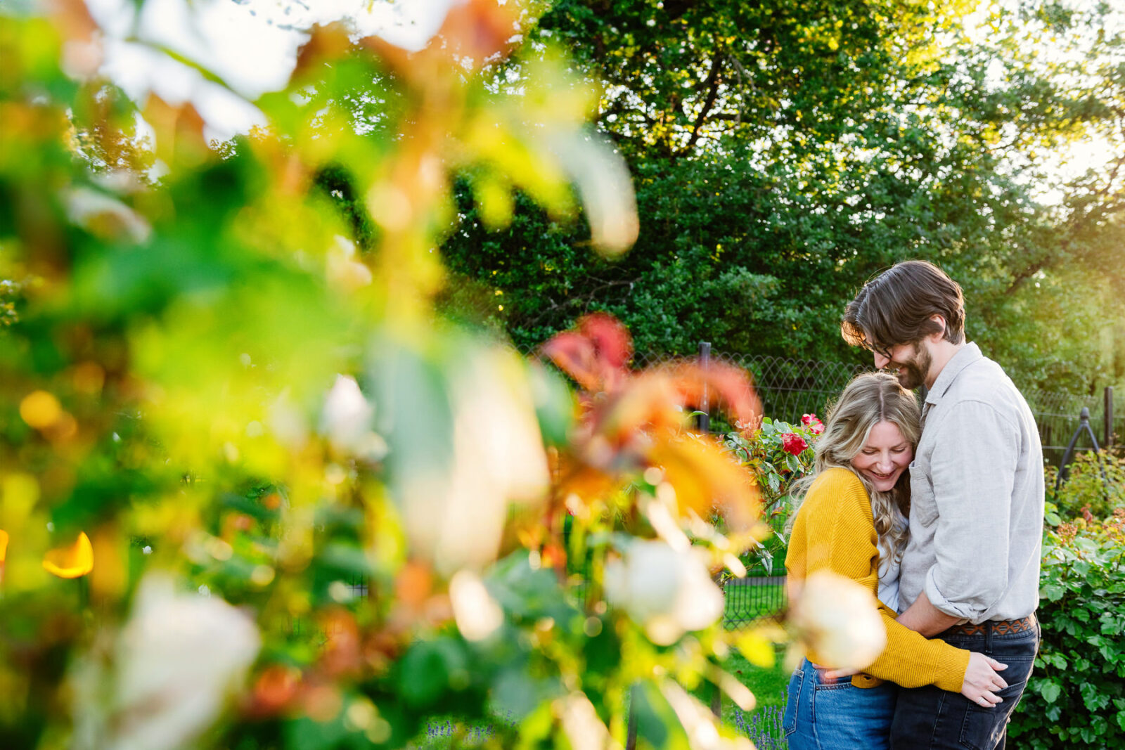 A couple snuggles with some leaves creating bokeh in the foreground during their engagement couples portrait session in Victoria BC