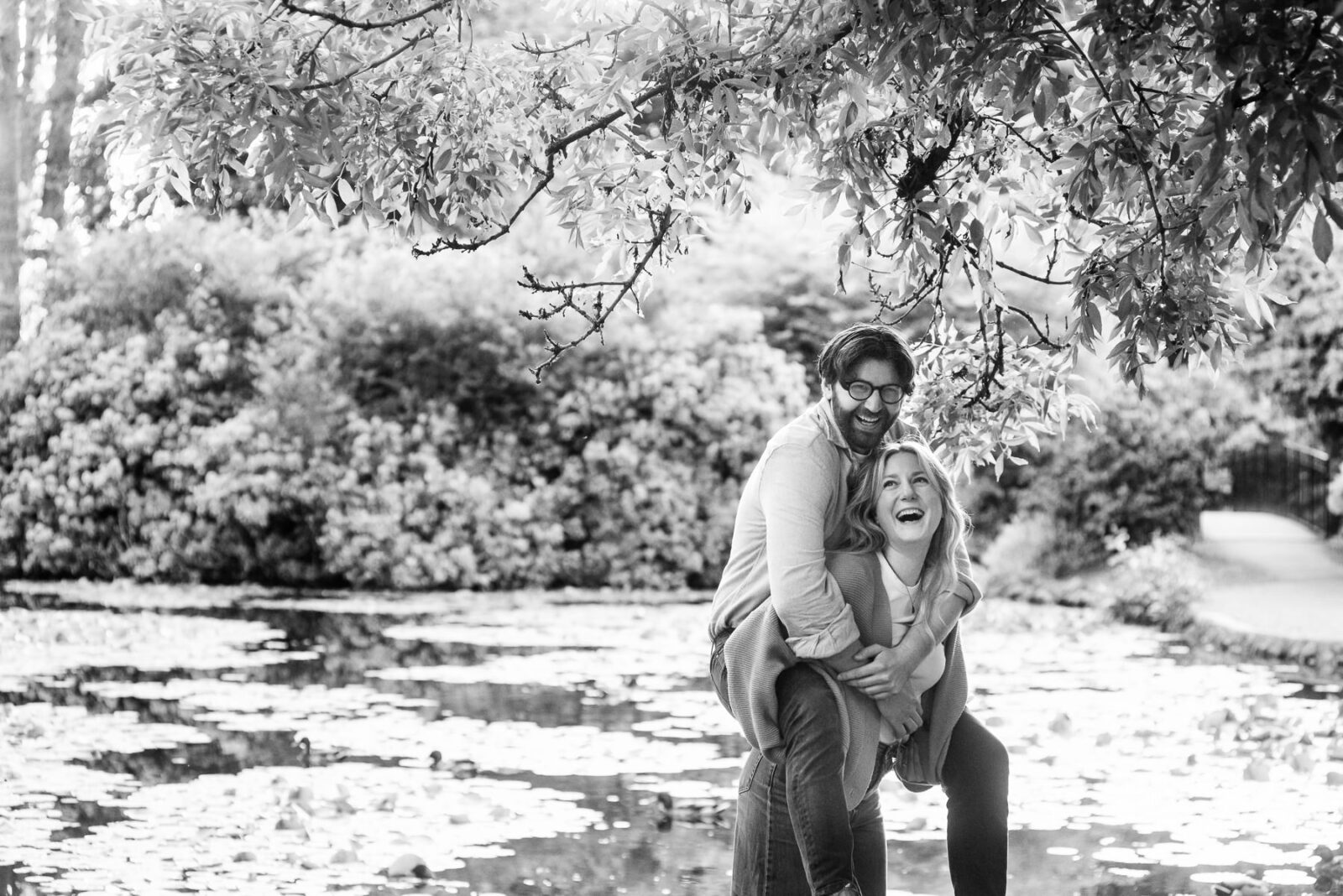 A guy gets a piggy back from his fiance in a black and white couples portrait taken at Beacon Hill Park in front of the lily pond.