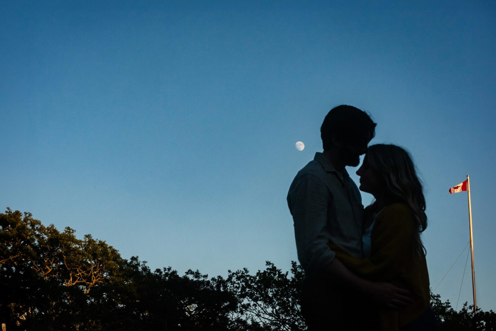 An evening engagement portrait of a couple standing in front of the moon and a Canadian flag. the couple is a silhouette.