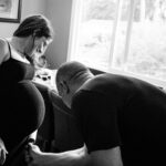 A father-to-be brushes off dog hair off mom-to-be's baby bump in their kitchen.