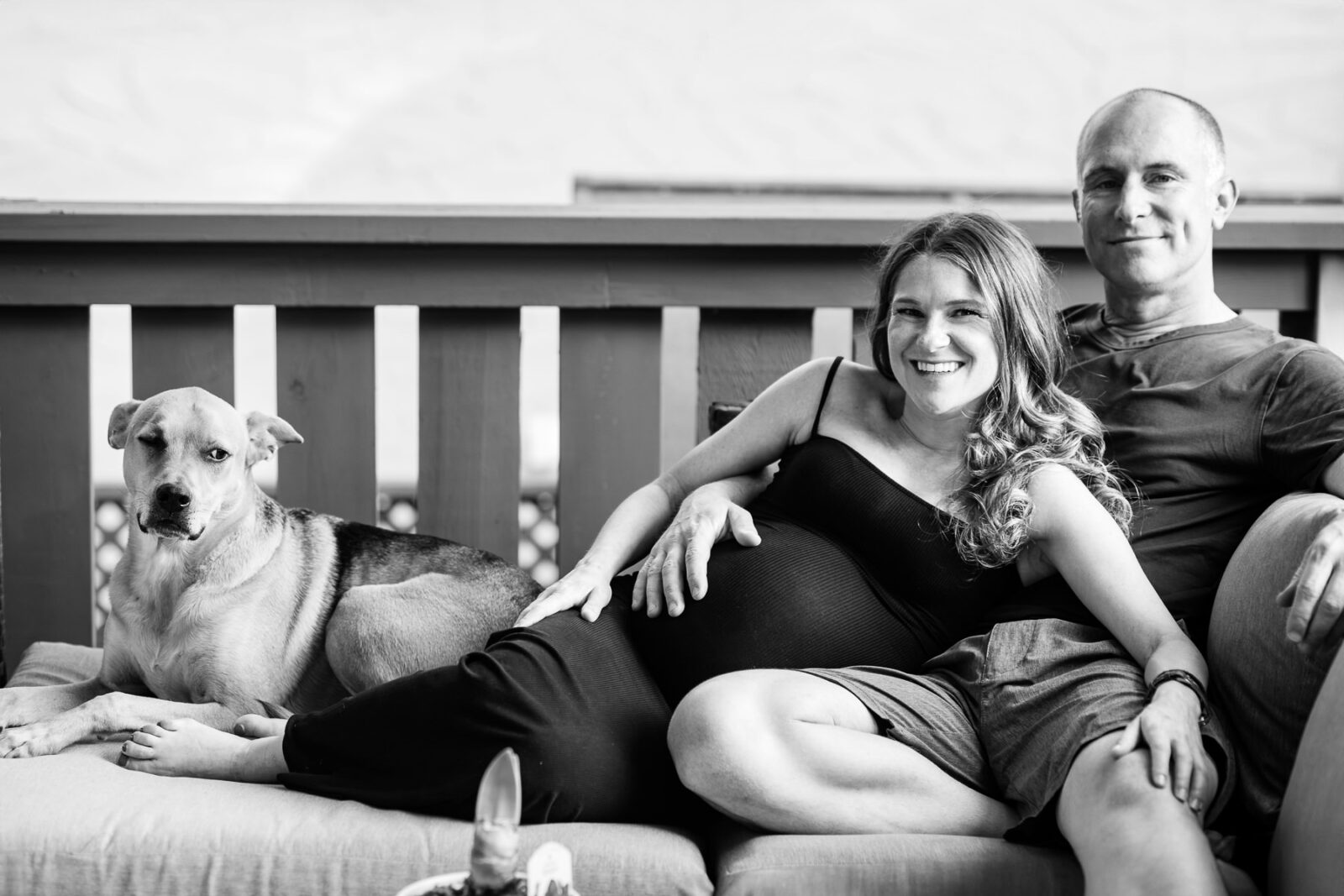 A couple stretches out on their patio couch with their dog. The father-to-be is holding his pregnant partner's baby bump during a relaxed maternity session at their home.