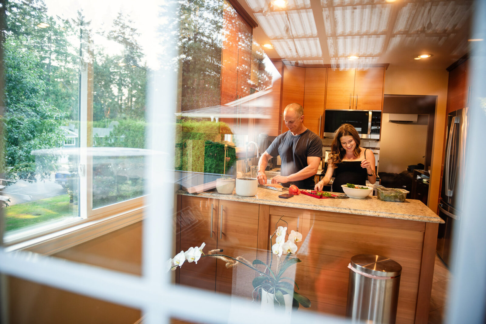A reflection through the french door window of a pregnant mom and her partner make dinner together during a candid and photojournalistic photo session at their house in Brentwood Bay.