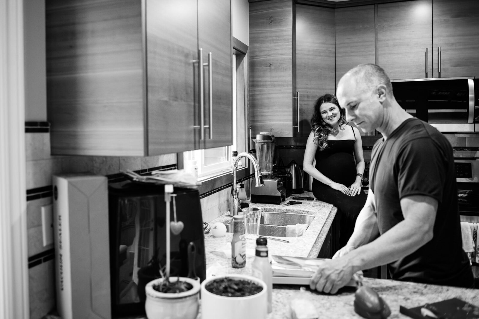 A pregnant mom sits on a counter while her partner make dinner together during a candid and photojournalistic photo session at their house in Brentwood Bay.