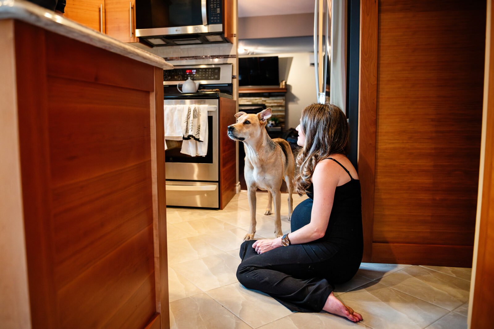 A pregnant mom-to-be laughing with her dog while sitting on the floor of her kitchen.