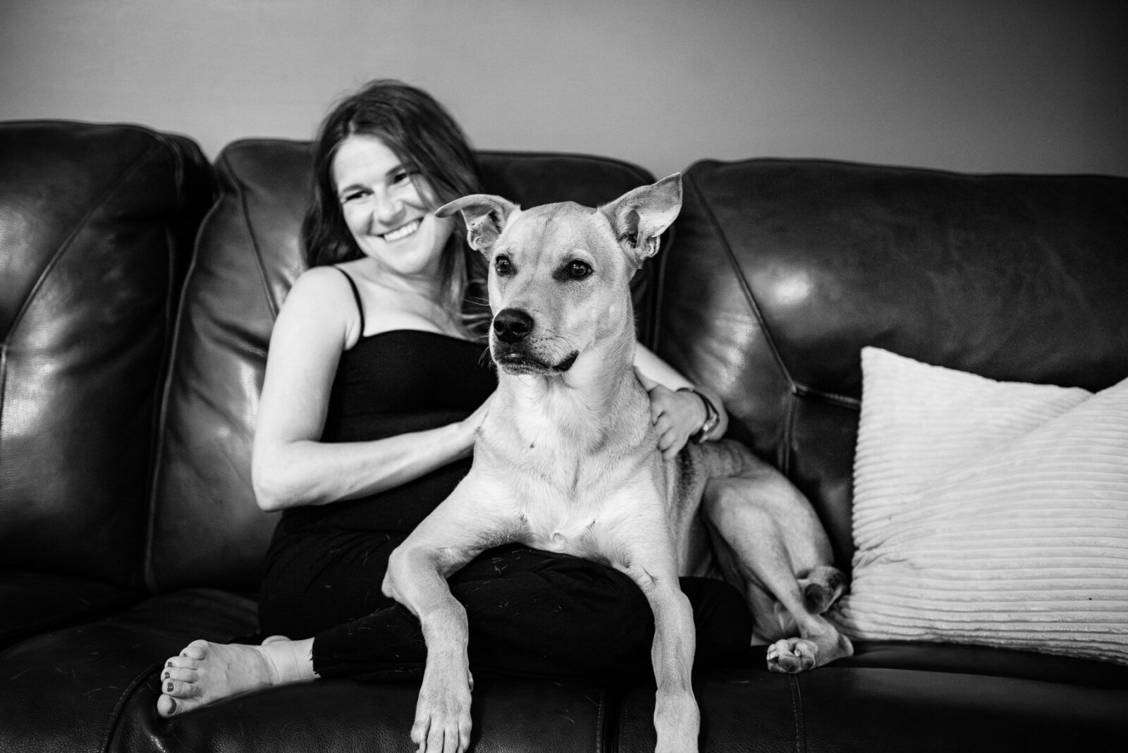 A mom to be laughs with her dog on her lap while sitting in their living room.