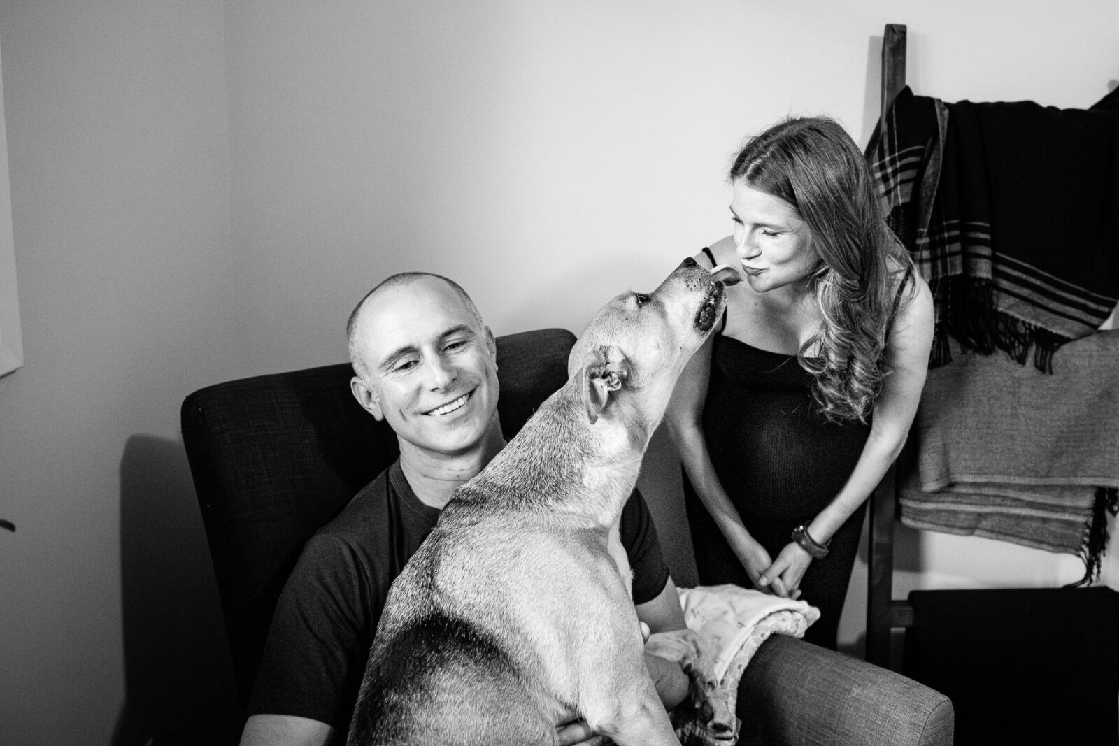 A mom-to-be kisses her dog while the father-to-be laughs.