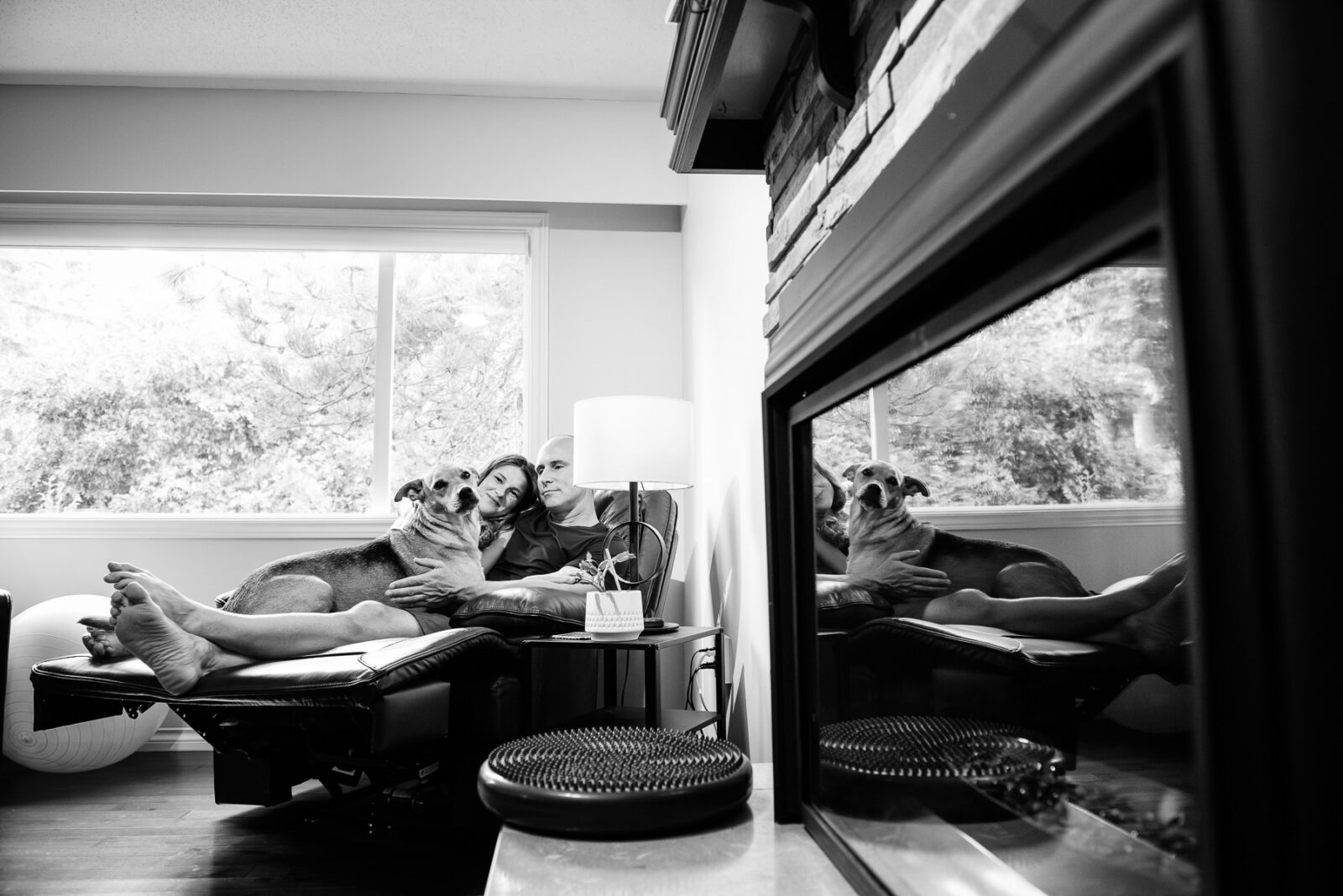 A couple kisses and hugs their dog in their family home with a reflection of them in the fireplace window.
