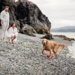 A couple laughs at their dog who has just come back from fetching a toy in the ocean at a VRBO rental in Sooke BC. This vacation portrait is candid and in colour.