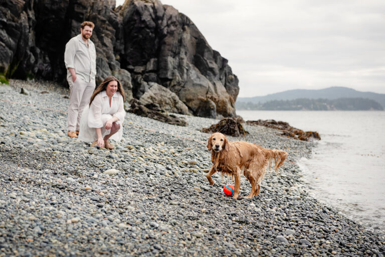 A couple laughs at their dog who has just come back from fetching a toy in the ocean at a VRBO rental in Sooke BC. This vacation portrait is candid and in colour.