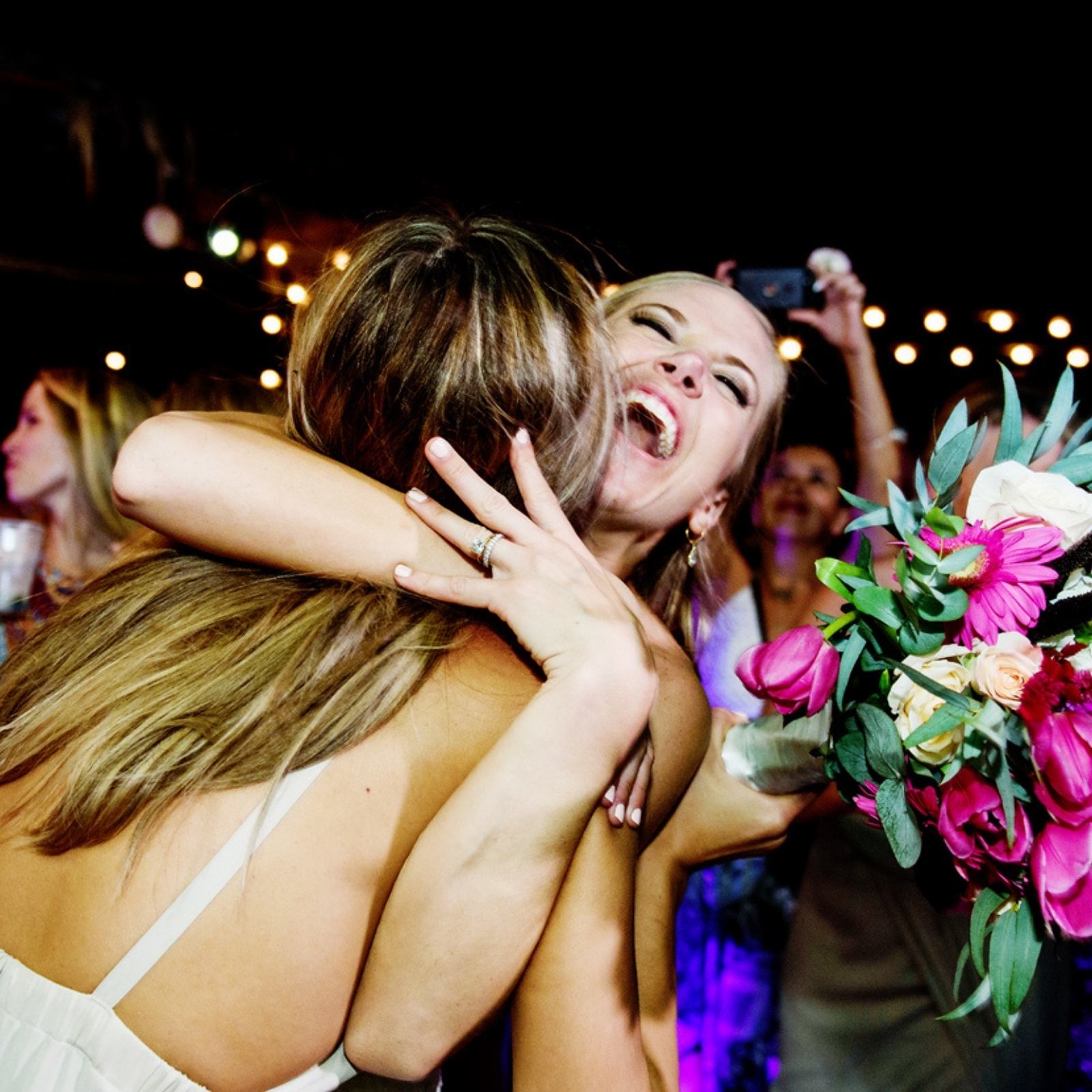 Brides hug each other while laughing on a wedding day in Victoria BC at night with people toasting in the background
