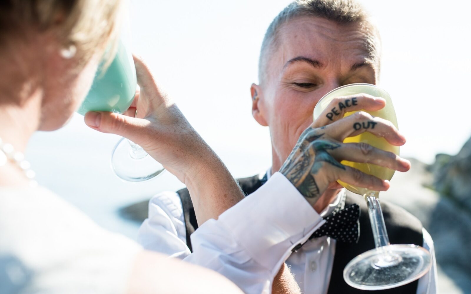 A trans couple links arms while they drink champagne after their outdoor wedding ceremony on Dallas Road in Victoria BC. One of the people has a tattoo that says peace out