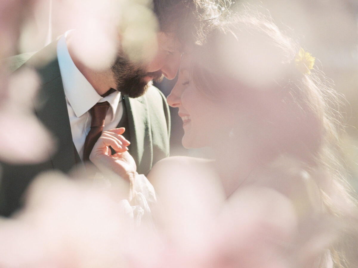 A soft dreamy film photo session of a bride laughing with her groom (a closeup) surrounded by cherry blossoms during an editorial photo session in Victoria BC