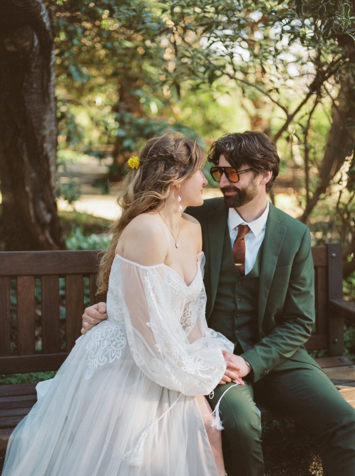 A bride wears a Persephone 50712 gown by Watters Designs purchased in Vancouver BC and the groom wears a green custom made Indochino suit. They are sitting on a park bench during an editorial photography session in Victoria BC