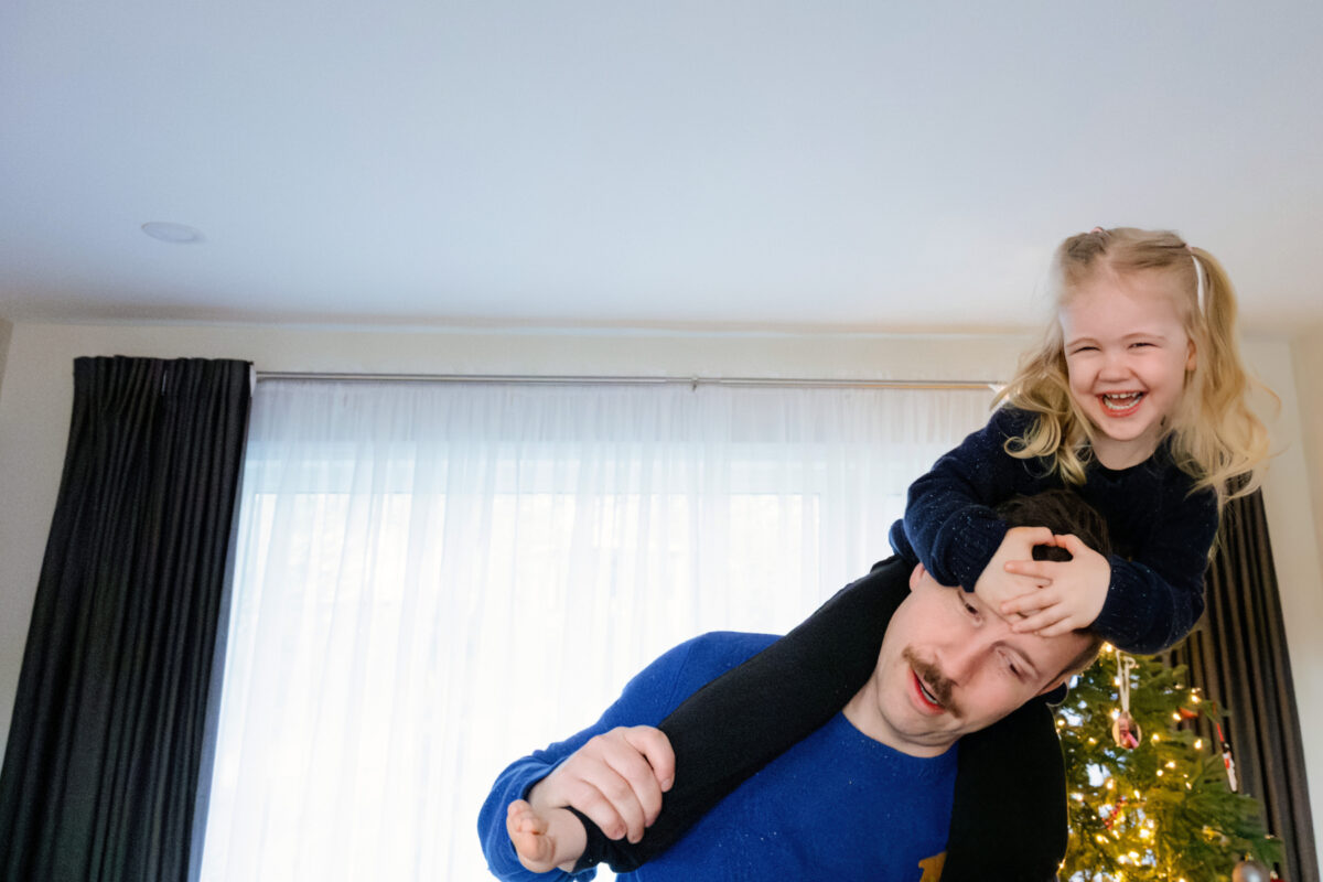 A funny photo of a child balancing on her father's shoulders in front of the christmas tree during a family mini session with candid photography in Victoria BC