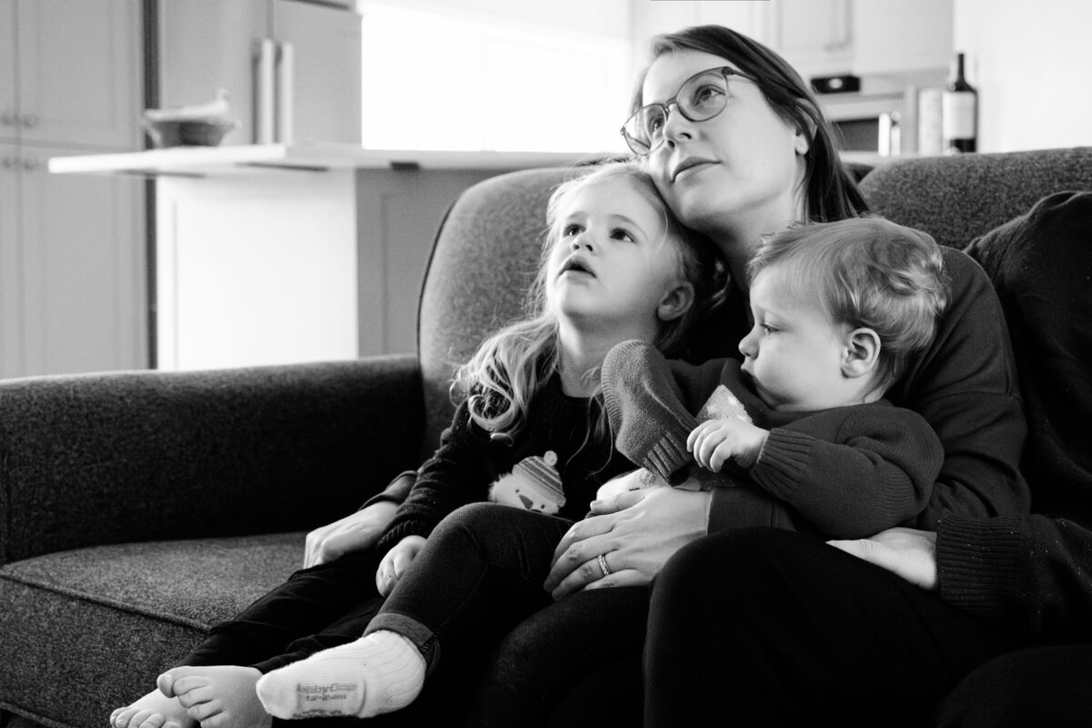 A mom cuddles with her toddlers while sitting on the couch watching TV during a documentary candid photo session in Victoria BC