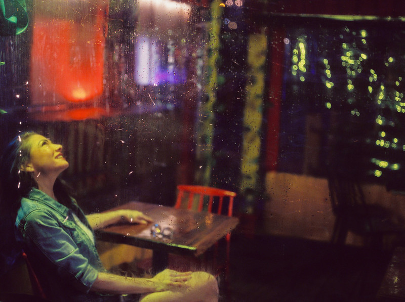 A cinematic portrait on Cinestill 800T film of a woman at night in an empty restaurant. She is looking up towards the light and there is a rainy tarp as a wall.