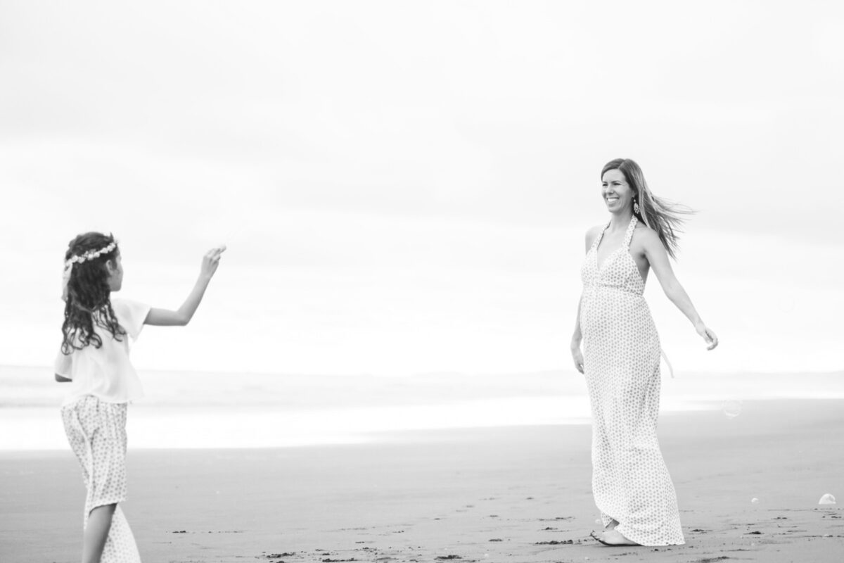 A big sister plays with bubbles while her pregnant mother twirls in a dress on the beach during a maternity mini-session at the ocean.
