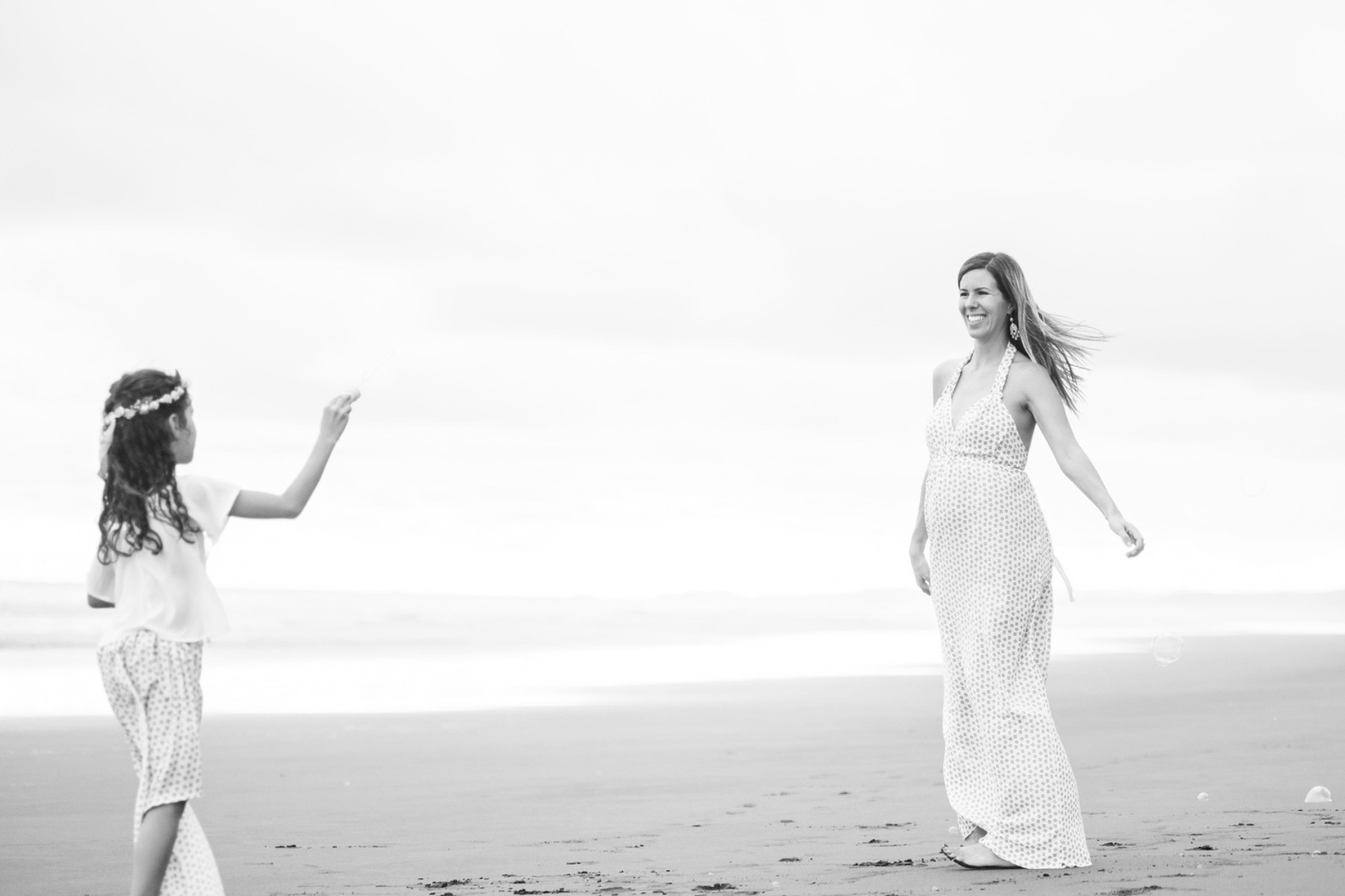A big sister plays with bubbles while her pregnant mother twirls in a dress on the beach during a maternity mini-session at the ocean.