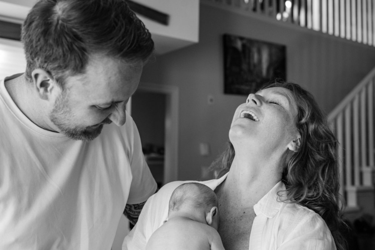 A candid documentary photojournalism image of parents holding their baby and laughing during an at-home newborn photo session in Langford BC