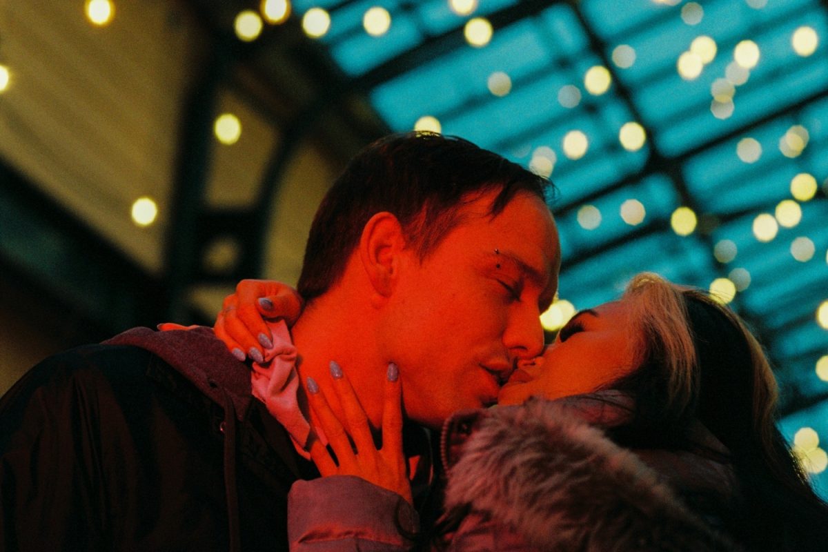 A sexy, grainy image of a couple kissing lit with a red filtered video light with bokeh in the background from the city lights in Victoria BC. shot on Kodak Ektacolor Pro 800 35mm film.