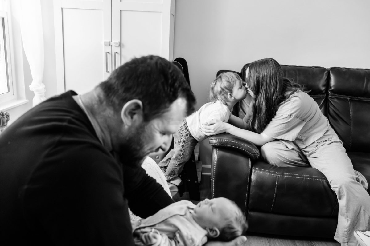 A toddler sibling kisses his mother while his father holds a newborn baby. The photo is in black and white and it was taken by a professional newborn and baby photographer in Victoria BC
