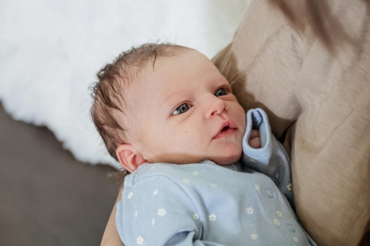 A colour photograph of a newborn baby being held by her mother. She is wearing a blue onesie and the photo is taken as part of natural, candid newborn photography in the family's home in Colwood near Victoria BC