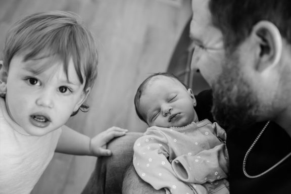 A new big brother sibling looks at the camera while his father holds his newborn baby sister during a family portrait session at home in Victoria BC