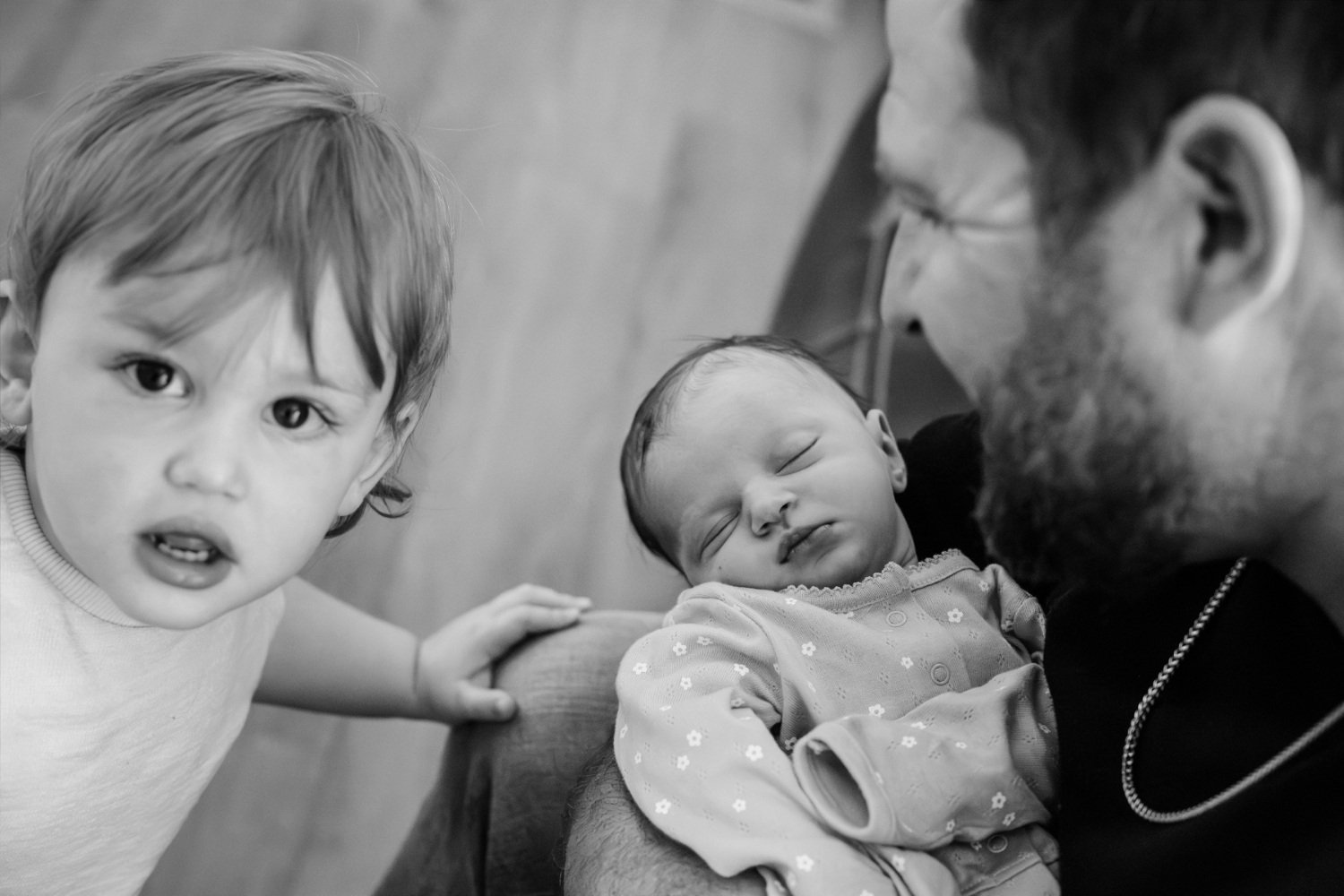 A new big brother sibling looks at the camera while his father holds his newborn baby sister during a family portrait session at home in Victoria BC
