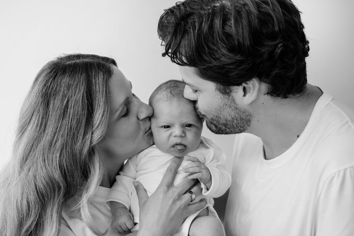 Newborn photo in black and white in Victoria BC at the couple's home. Mom and dad are kissing their baby on each side.