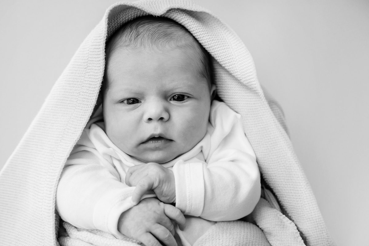 An at-home newborn mini-session in the family home in black and white. A baby looks at the camera with a blanket around her head.