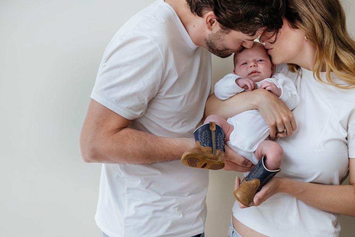 Mom and dad are kissing their baby during a candid moment at their home for a newborn photography session. The baby is wearing cowboy boots.