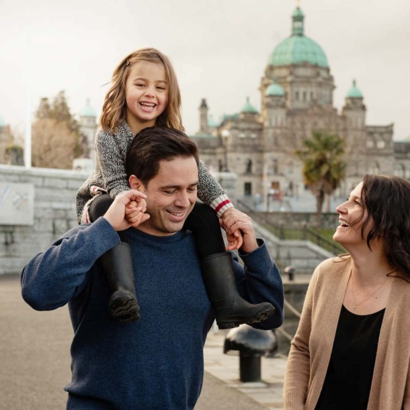 A family of three with a toddler having a fun time with a child on dad's shoulders in front of the BC Legislature buildings in Victoria BC. The photo is in colour and this is a candid family portrait.