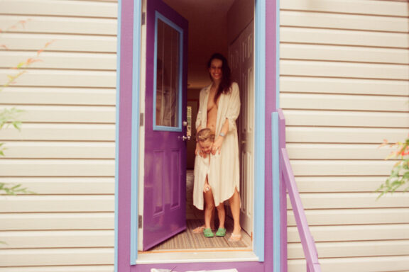 A blurry film photo taken in 2026 using Kodak Vision 3 motion picture stock. A mother his holding her child in front of their purple cottage door.