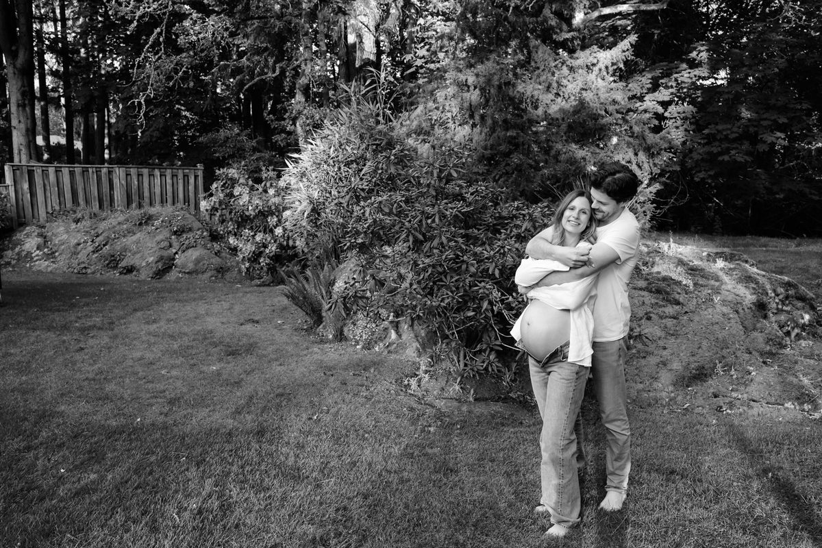 A black and white maternity portrait of a couple hugging outside with the belly exposed. The mother is wearing a white shirt and jeans.